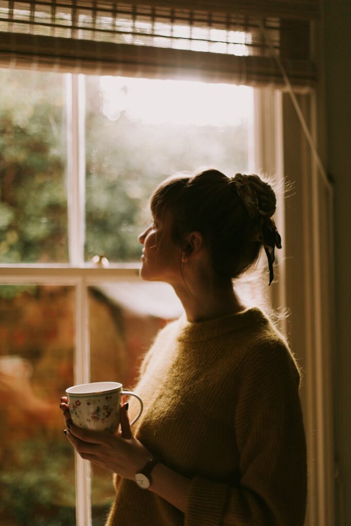 Une femme tient une tasse près d’une fenêtre, dans une lumière douce